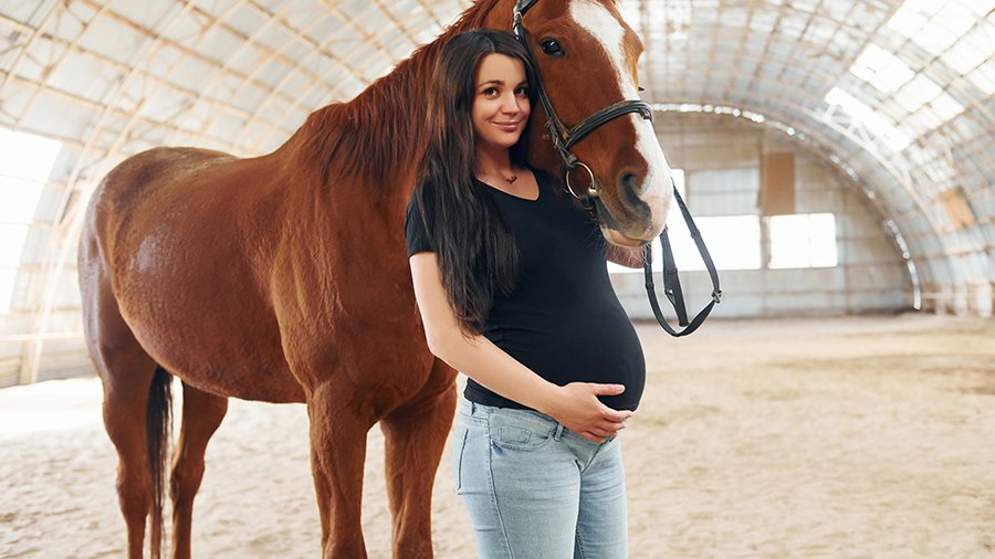 Schwangere Frau mit dunkelbraunen langen Haaren in Jeans und schwarzem Shirt steht neben braunem Pferd mit großer weißer Blesse in einer hellen Reithalle und schaut in die Kamera.