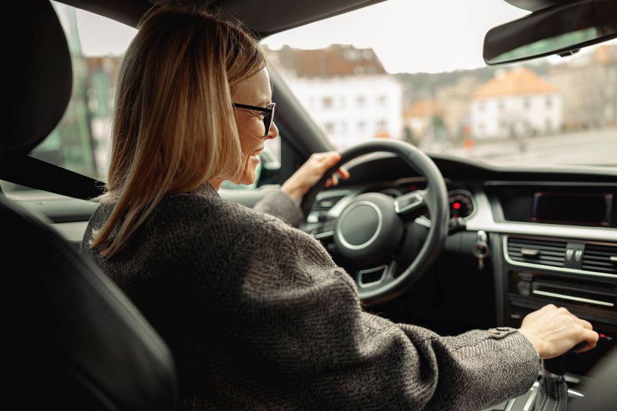 Blick vom Rücksitz eines Autos auf blonde Fahrerin, die aus dem Fenster schaut und lacht, eine Hand am Lenkrad, eine am Schalthebel. Kleidung und Fahrzeuginneres überwiegend grau.