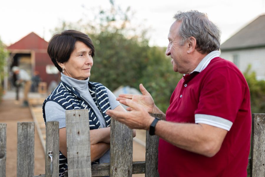 Gespräch am Zaun, Frau mittleren Alters hört freundlich und aufmerksam zu, Mann mit rotem Poloshirt etwas älter gestikuliert und redet auf sie ein.