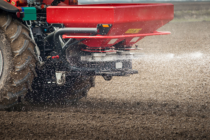 Traktor mit Düngerstreuer bringt granulierten, weißen Dünger auf einen unbewachsenen Acker aus.