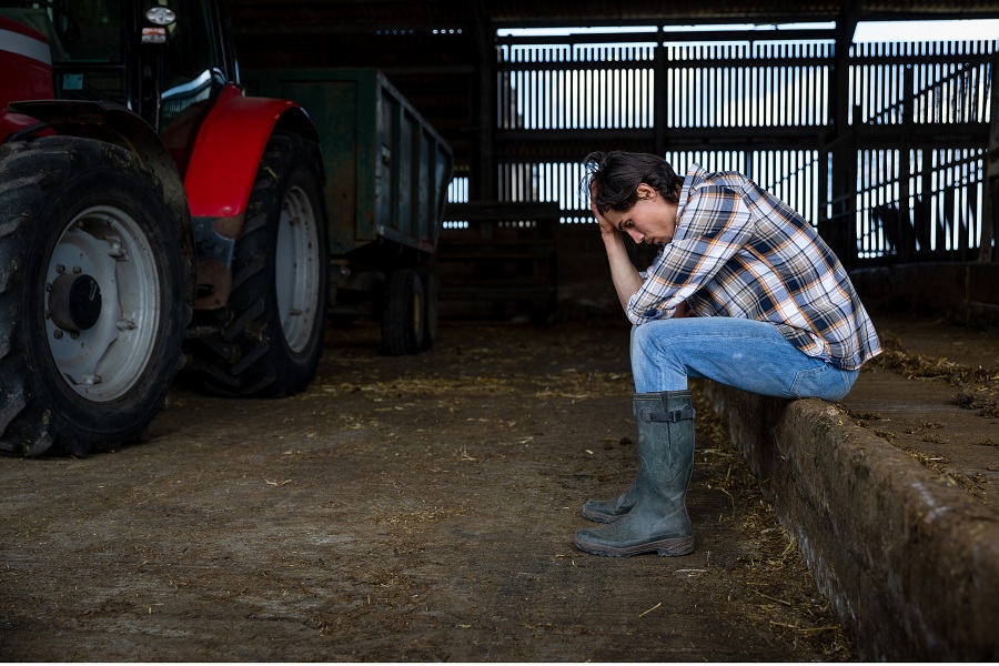 Junge Bäuerin sitzt neben einem Traktor und stützt den Kopf ab.