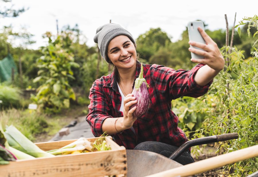 Frau hockt in einem Gemüsefeld und macht ein Selfie. Sie hält eine Aubergine in der einen und ein Handy in der anderen Hand.