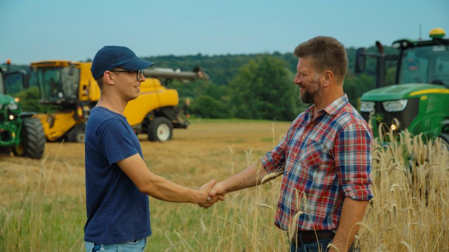 Zwei Männer stehen auf einem Feld und geben sich die Hand. Im Hintergrund sieht man landwirtschaftliche Fahrzeuge.