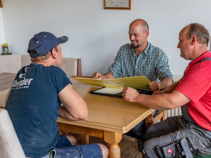 Drei Männer sitzen an einem Tisch, einer hält einen geöffneten gelben Ordner in der Hand. Er lacht. Die anderen beiden tragen typische landwirtschaftliche Arbeitskleidung.