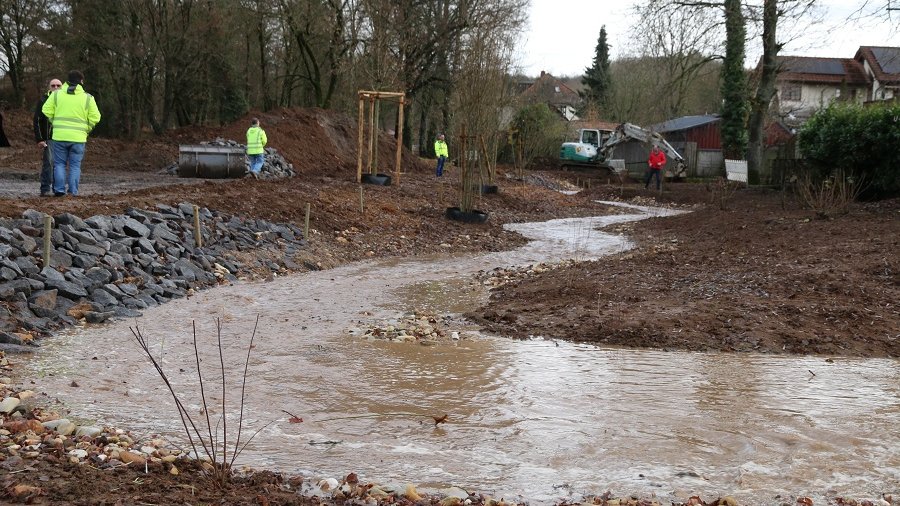 Ein Bach windet sich durch eine Baustelle. Am Rand werden Steine verlegt und es ist viel Erde zu sehen.