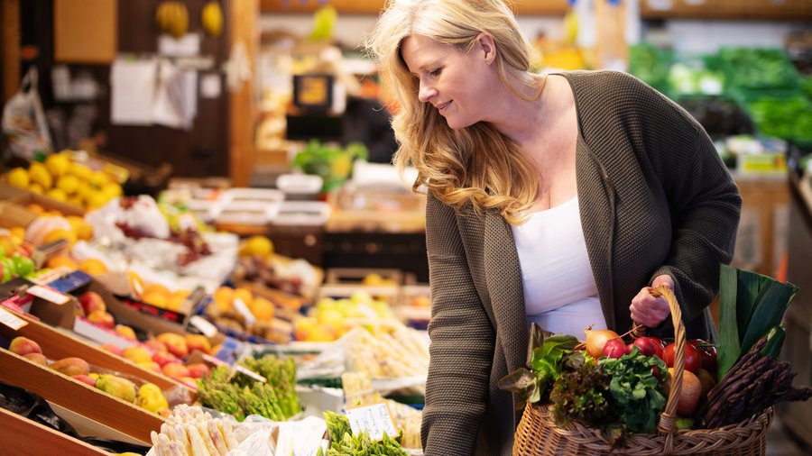 Eine Frau beugt sich in einem Hofladen über Kisten mit Obst und Gemüse und schaut diese an, dabei hält sie einen Korb mit Produkten in der Hand. 