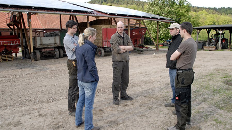Vier Landwirte und eine Landwirtin stehen auf Hofgelände und unterhalten sich. Im Hintergrund ist eine große Maschinenhalle zu sehen.