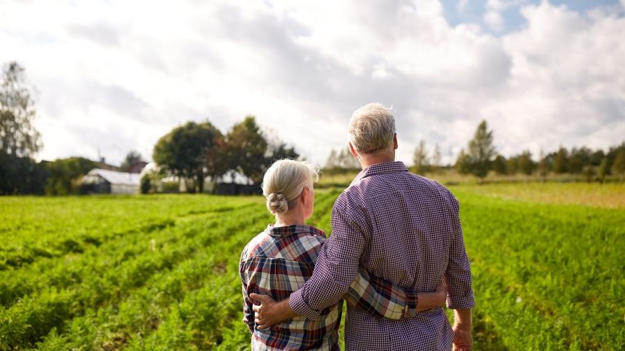 Ältere Frau und älterer Mann mit Rücken zur Kamera halten über Kreuz die Arme um die Taille des anderen und schauen über grüne Felder. Im Hintergrund Bäume und weißblauer Himmel.