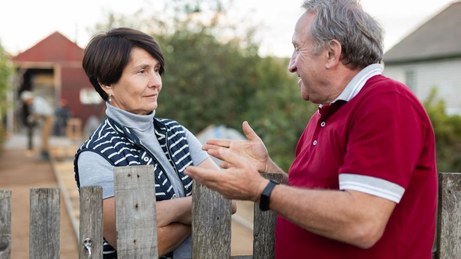 Gespräch am Zaun, Frau mittleren Alters hört freundlich und aufmerksam zu, Mann mit rotem Poloshirt etwas älter gestikuliert und redet auf sie ein.