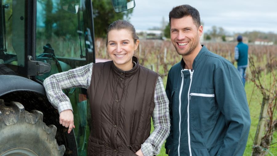 Junge Frau und junger Mann stehen in einer Obstplantage neben Schlepper und lachen in die Kamera. 