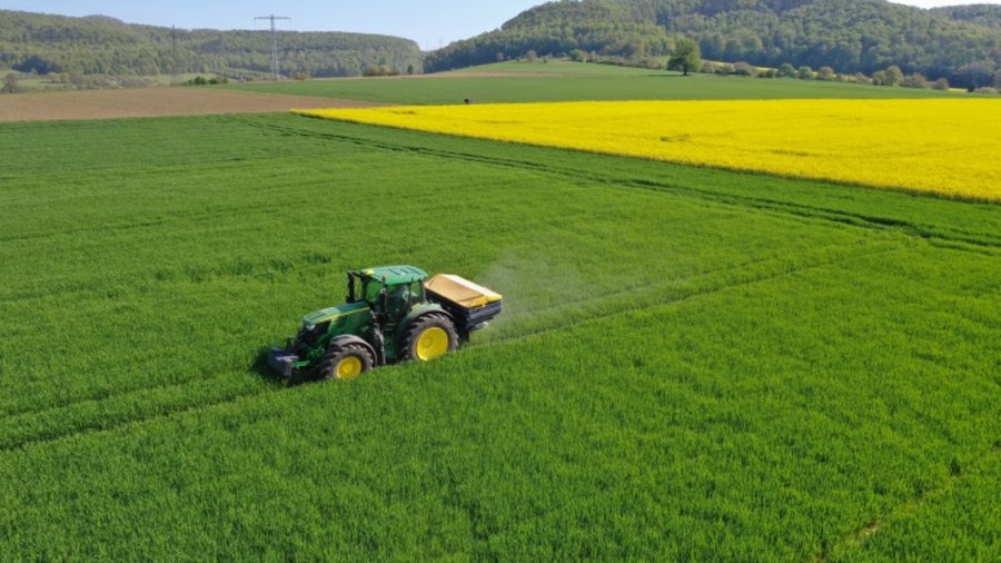  Ein Schlepper mit Düngerstreuer bei einer Stickstoffgabe im Winterweizen. Im Hintergrund ein blühendes Rapsfeld.