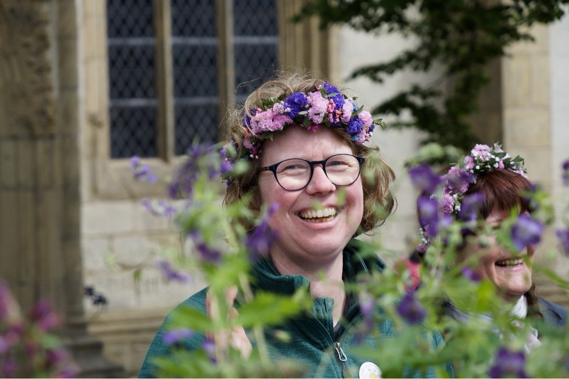 Zu sehen ist ein Portrait von Christiane Kalbe mit einem Blumenkranz auf dem Kopf. Sie schaut zwischen Pflanzen hindurch.