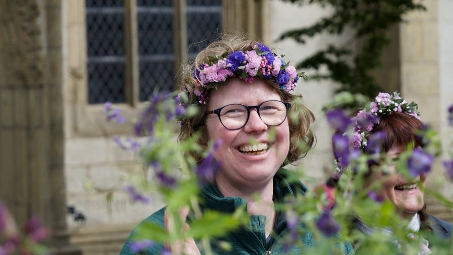Zu sehen ist ein Portrait von Christiane Kalbe mit einem Blumenkranz auf dem Kopf. Sie schaut zwischen Pflanzen hindurch.