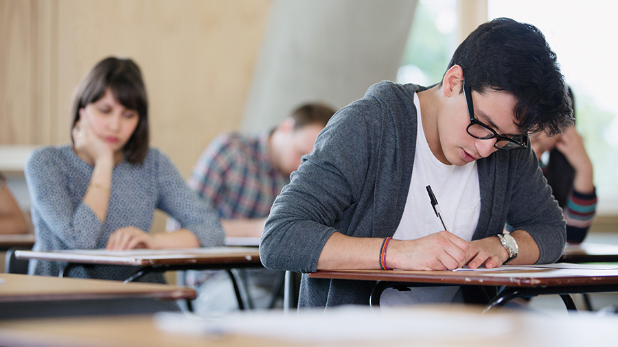 Junger Mann und junge Frau sitzen an Tischen und schreiben angestrengt einen Test.