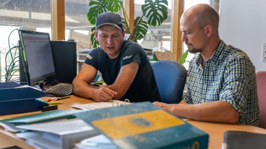 Zwei Männer sitzen nebeneinander an einem Schreibtisch mit Akten und Computerbildschirm. Sie schauen in Unterlagen, die vor ihnen auf dem Tisch liegen. Im Hintergrund ist ein großes mehrteiliges Fenster zum Bauernhof zu sehen.