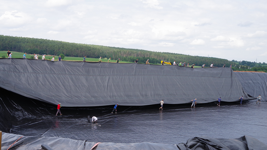 Zu sehen sind Personen, die am Rande des Beckens stehen und die schwarze Folie systematisch ausbringen.