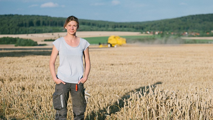 Junge Frau steht auf einem Weizenfeld. Im Hintergrund sieht man einen Mähdrescher.