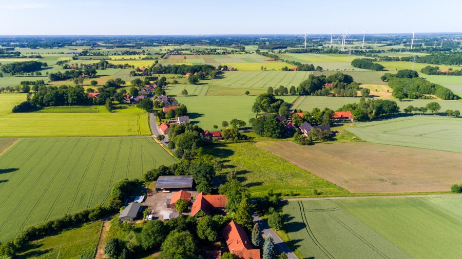 Luftaufnahme einer landwirtschaftlich geprägten Region mit einem Betrieb, Feldern und Windrädern im Hintergrund.