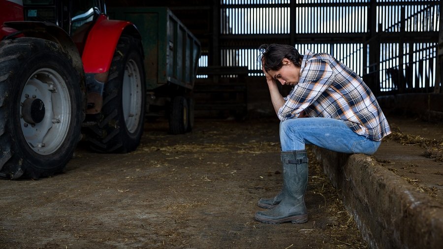 Junge Bäuerin sitzt neben einem Traktor und stützt den Kopf ab.
