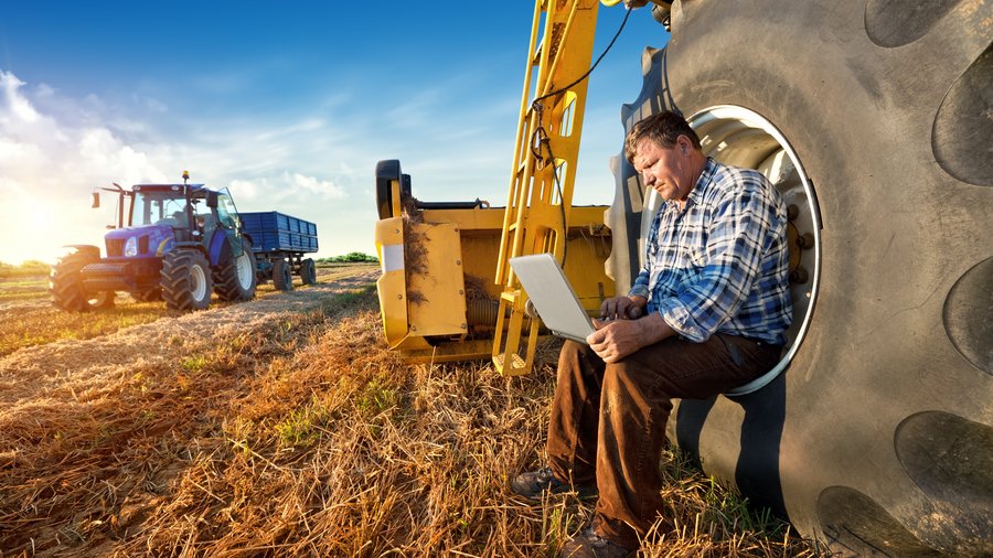 Ein Landwirt sitzt mit seinem Laptop an einem Traktor auf dem Feld.