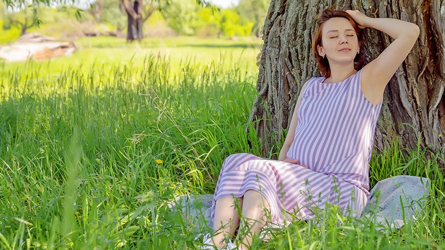 Schwangere Frau sitzt an einen alten Baum gelehnt im Schatten, trägt ein längsgestreiftes weites Kleid. Sie hat die Augen geschlossen und erholt sich. Um sie herum steht hohes leuchtend grünes Gras, im Hintergrund ist gemähter Rasen und ein Baum verschwommen erkennbar.