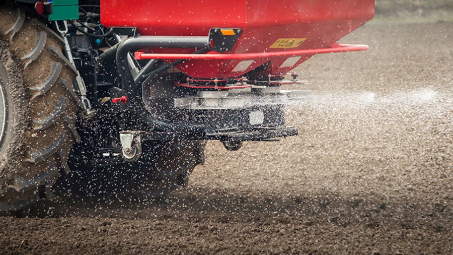 Traktor mit Düngerstreuer bringt granulierten, weißen Dünger auf einen unbewachsenen Acker aus.