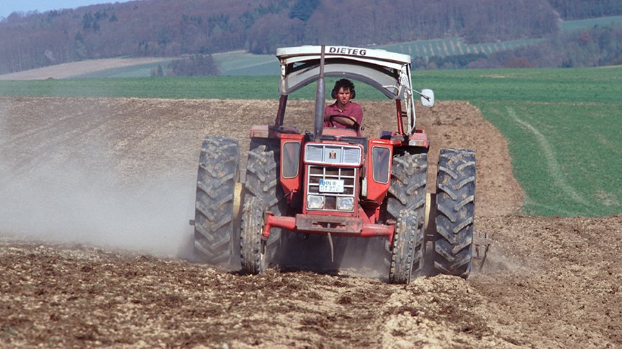 Roter Schlepper mit Zwillingsbereifung beim Grubbern im Frühjahr fährt auf Fotografen zu. Durch die geöffnete Frontscheibe sieht man Fahrer mit Ohrenschützern.