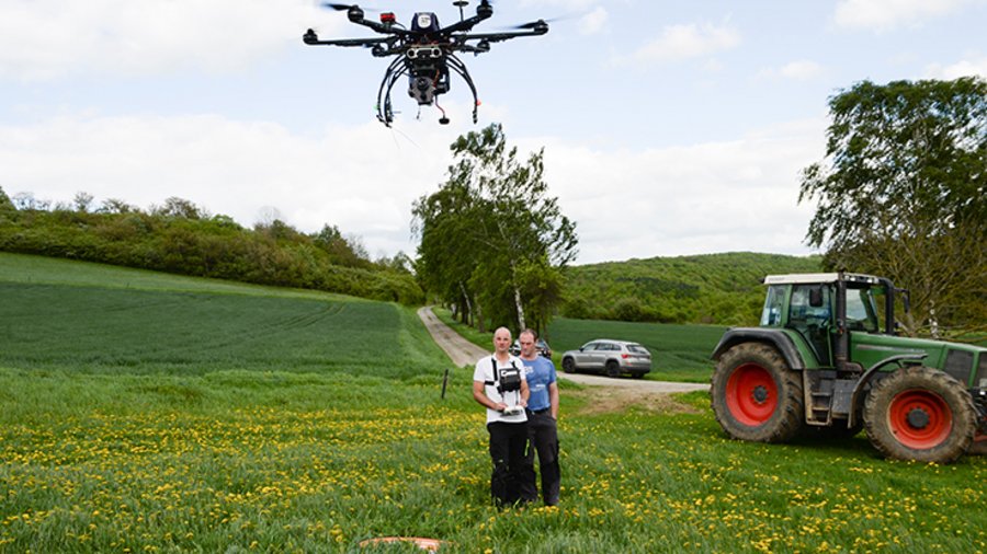 Im Vordergrund fliegt eine Drohne. Im Hintergrund stehen zwei Männer auf einer Wiese. Einer der Männer steuert die Drohne.