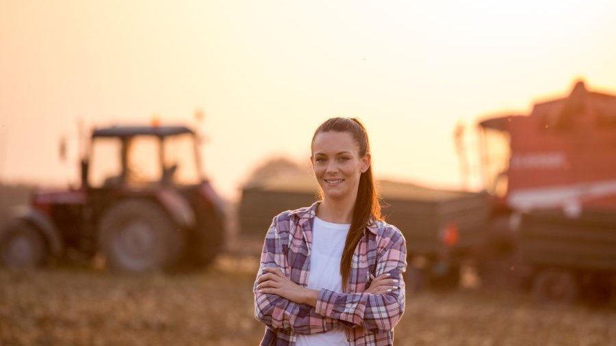 Funge Frau steht auf einem Feld, im Hintergrund sieht man Landmaschinen. Bild: Budimir Jevtic - stock.adobe.com 