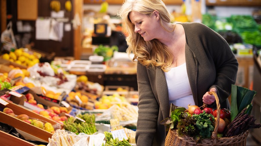 Eine Frau beugt sich in einem Hofladen über Kisten mit Obst und Gemüse und schaut diese an, dabei hält sie einen Korb mit Produkten in der Hand. 