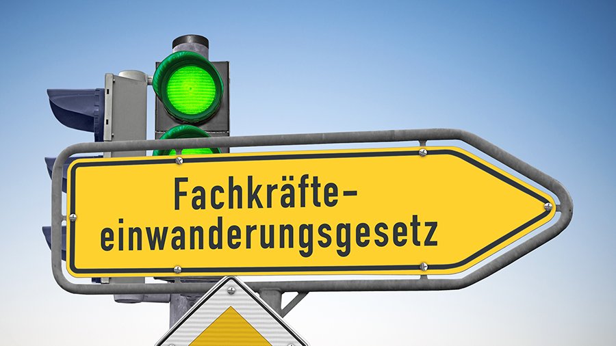 Gelbes richtungsangebendes Verkehrsschild mit Aufschrift „Fachkräfteeinwanderungsgesetz“, im Hintergrund Ampel, die ganz auf grün steht, im Vordergrund gelb-weißes Vorfahrtsstraßen-Schild.