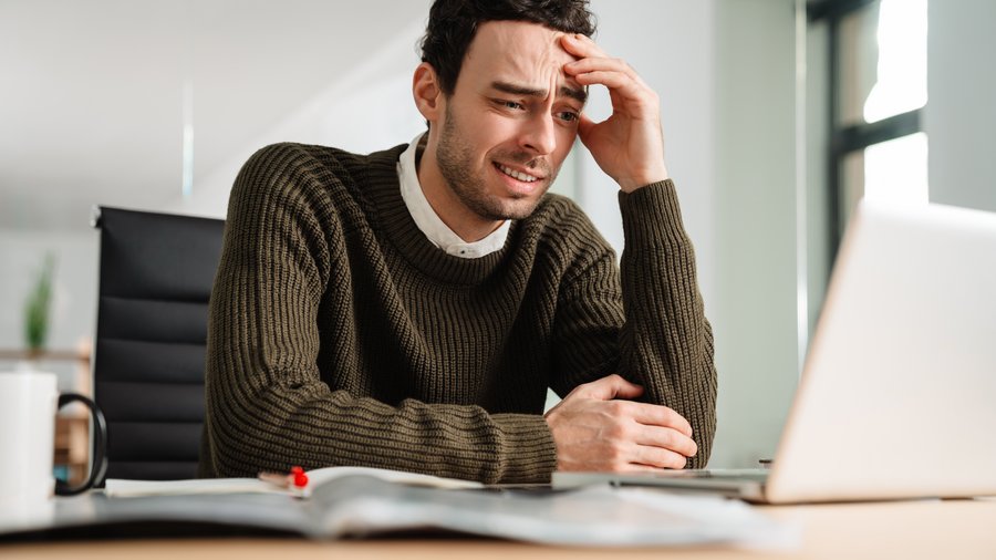 Ein Mann in braunem Pullover blickt verwirrt auf einen Laptop und greift sich an die Stirn.