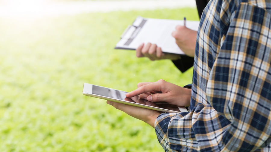 Zwei Personen stehen nebeneinander, seitlich aufgenommen, die vordere mit blau-weiß-gelb-kariertem Hemd. Eine Person hält ein Tablet in der Hand, die andere Person hält einen Notizblock auf einem Klemmbrett und schreibt darauf.