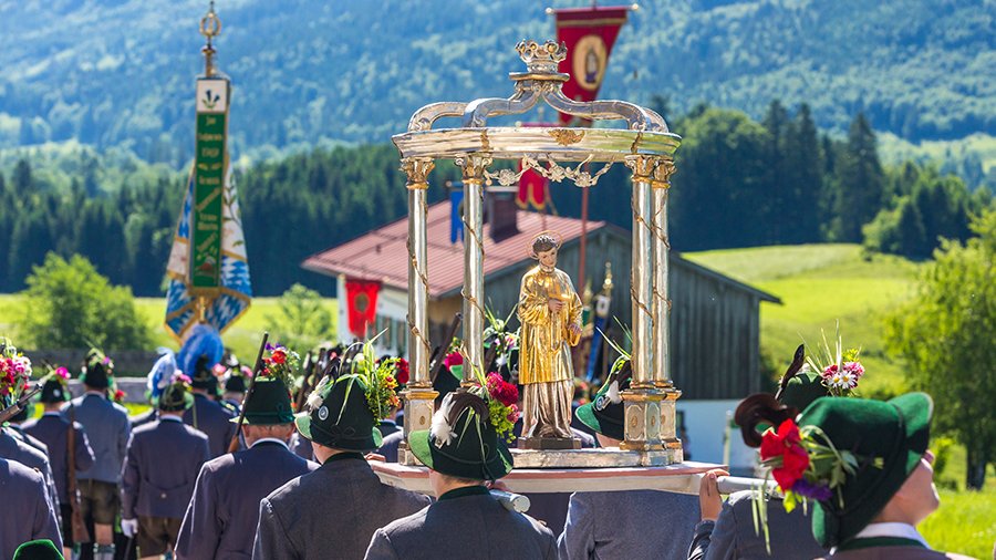 Männer in Trachten gehen von Kamera weg, tragen an Fronleichnam eine Jesus-Statue.