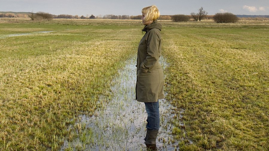 Frau in Gummistiefeln steht in überflutetem Teil einer Wiese.