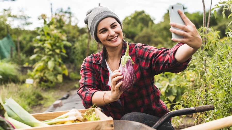 Frau hockt in einem Gemüsefeld und macht ein Selfie. Sie hält eine Aubergine in der einen und ein Handy in der anderen Hand.