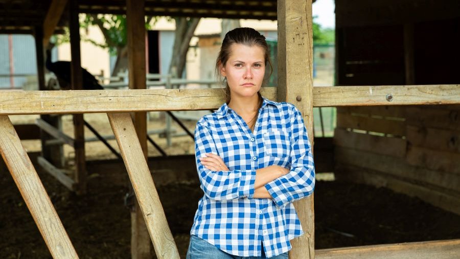 Frau mit blau-weiß kariertem Hemd und verschränkten Armen und traurigem und missmutigem Blick steht an Holzgatter eines überdachten Stalls. Hinter ihr seht man den leeren Stall.