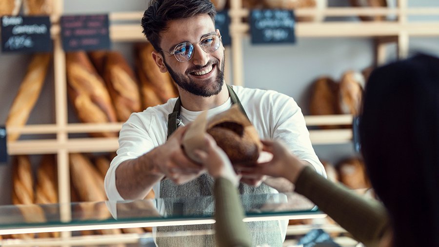 Ein junger Verkäufer reicht einer Kundin ein Brot über die Theke, das lose in Papier eingeschlagen ist. 
