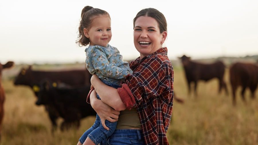 Lachende Frau mit rot-kariertem Hemd und etwa 2- bis 3-jährigem Mädchen auf dem Arm schaut in Kamera, von der Seite fotografiert, im Hintergrund verschwommen Rinder auf Weide mit braunem Gras.