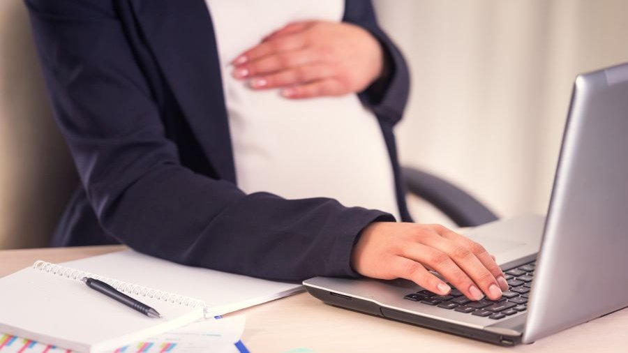 Frau mit dunkelgrauer Jacke und weißem Shirt sitzt vor Laptop, eine Hand auf der Tastatur, eine auf dem schwangeren Bauch liegend, Kopf nicht sichtbar. Auf dem Tisch liegen weitere Schreibutensilien.