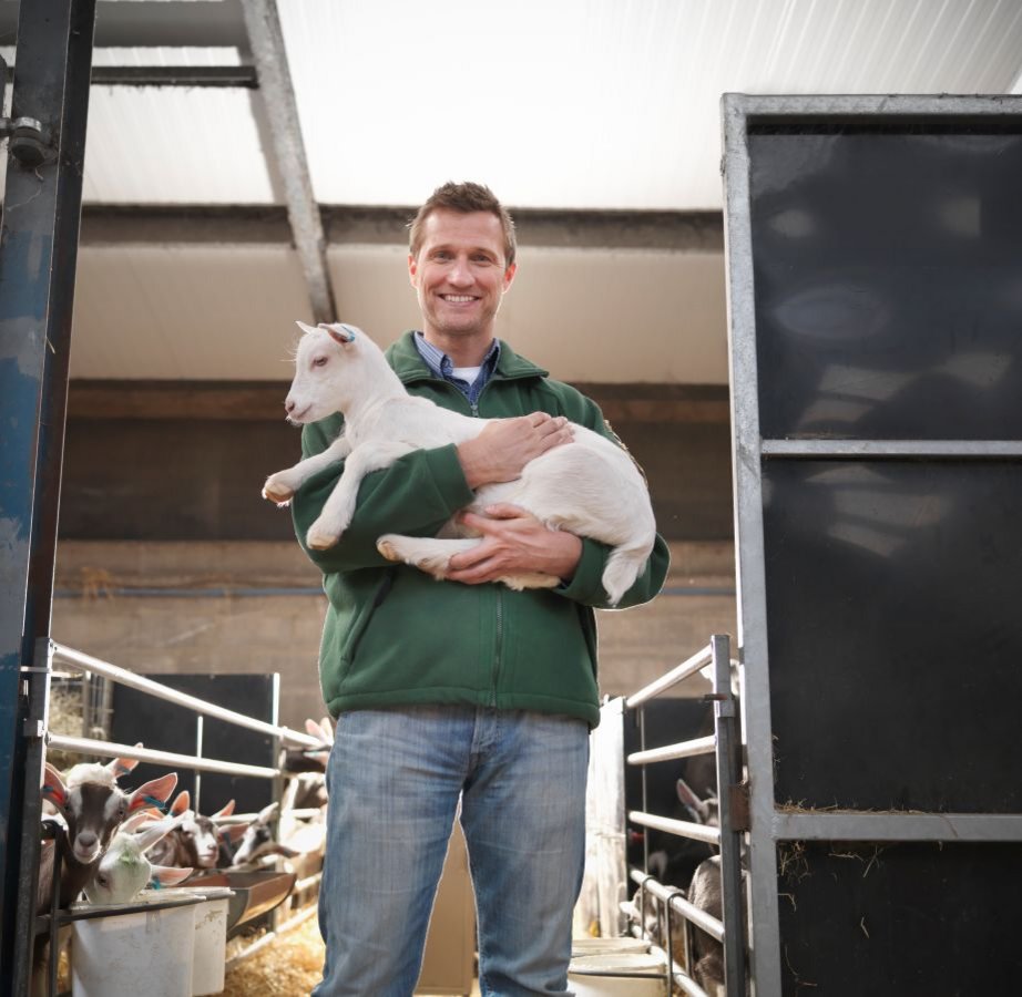 Mann mit Zicklein auf dem Arm steht im Stall. Klick öffnet Großansicht im neuen Fenster.