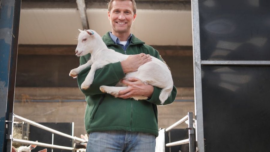 Mann mit Zicklein auf dem Arm steht im Stall. Klick öffnet Großansicht im neuen Fenster.