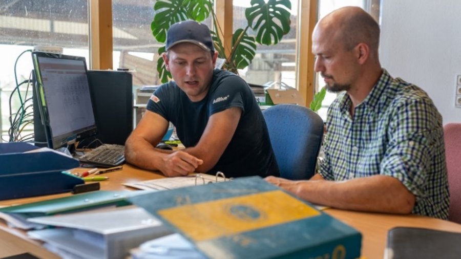 Zwei Männer sitzen nebeneinander an einem Schreibtisch mit Akten und Computerbildschirm. Sie schauen in Unterlagen, die vor ihnen auf dem Tisch liegen. Im Hintergrund ist ein großes mehrteiliges Fenster zum Bauernhof zu sehen.
