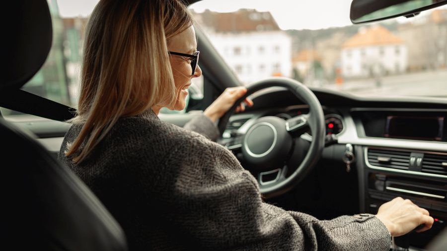 Blick vom Rücksitz eines Autos auf blonde Fahrerin, die aus dem Fenster schaut und lacht, eine Hand am Lenkrad, eine am Schalthebel. Kleidung und Fahrzeuginneres überwiegend grau.